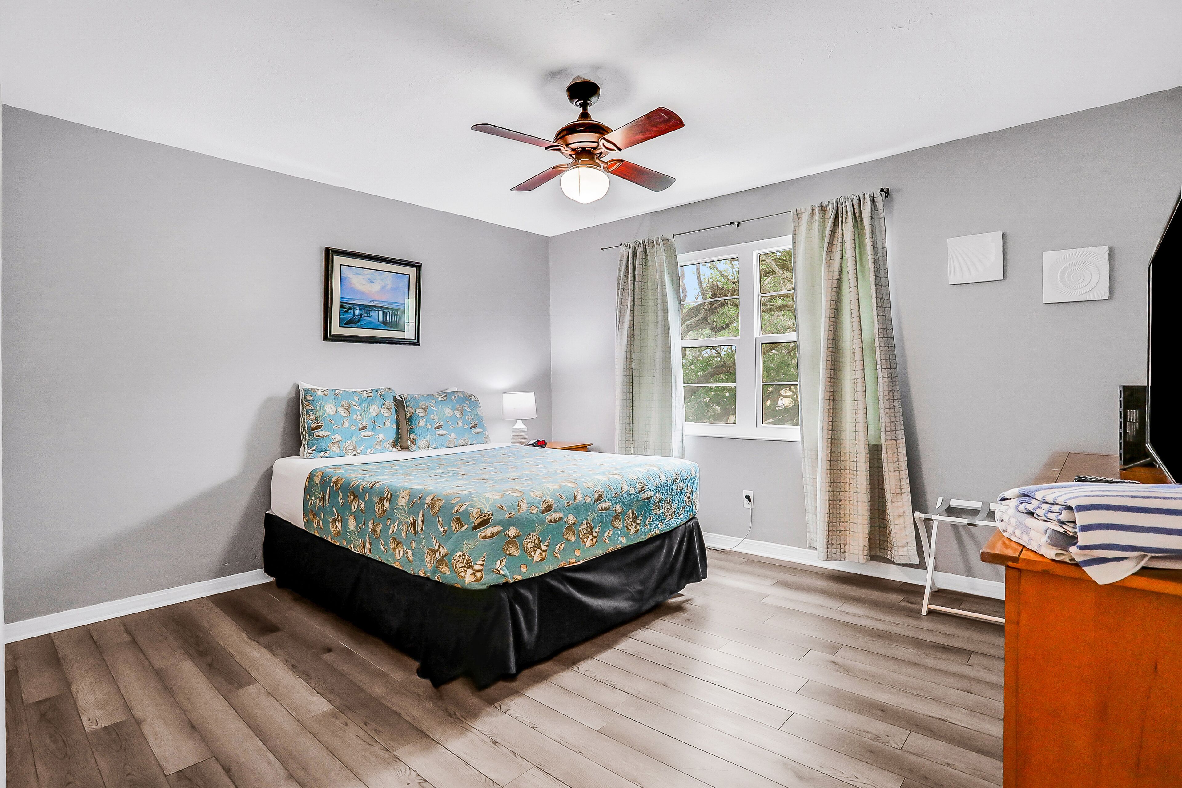 A bedroom with a bed, a ceiling fan, a window with curtains, a bedside table, and a framed picture on the wall.
