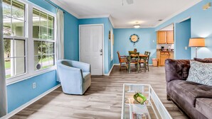 A living room with a blue wall, a ceiling fan, a dining area with a table and chairs, and a kitchen area with cabinets and a lamp. - Charming Condo w/ Pool 500 Yards to Beach, Near Lighthouse & Downtown (St. Augustine)