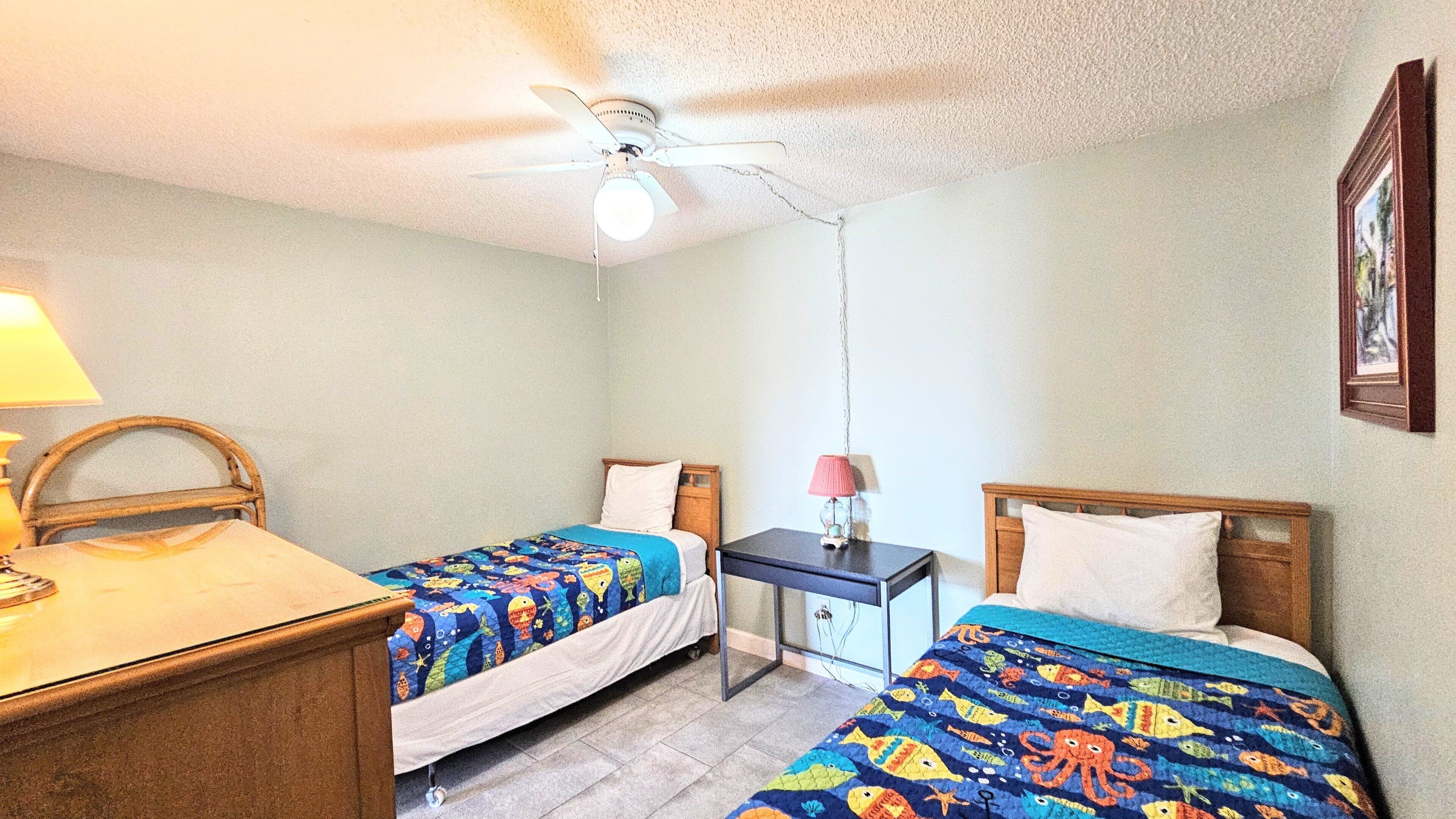 A room with two beds, a desk, a chair, a ceiling fan, and a small table with a lamp.
