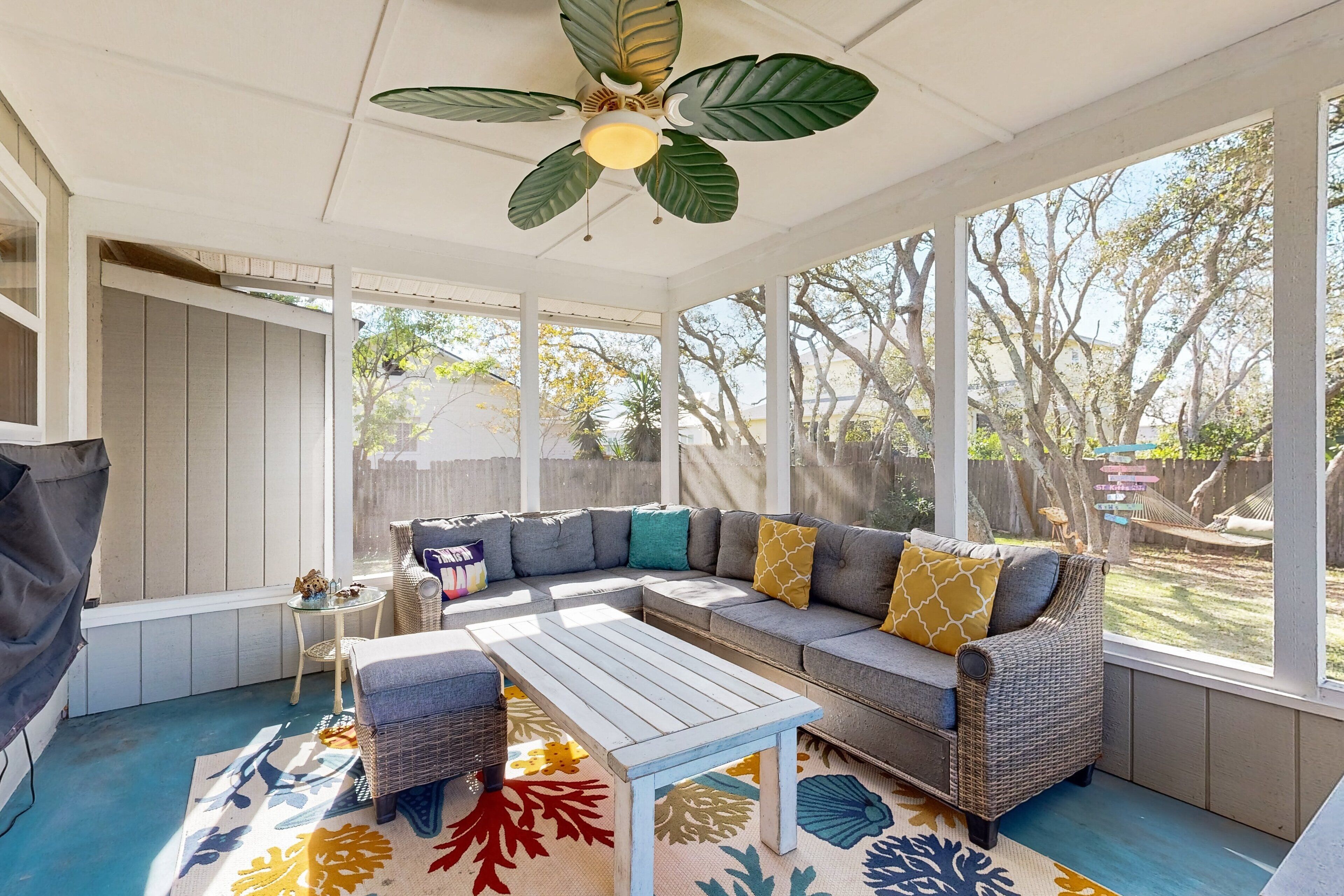 Une terrasse couverte avec un ventilateur de plafond, un canapé d’angle, une petite table et un tapis coloré.