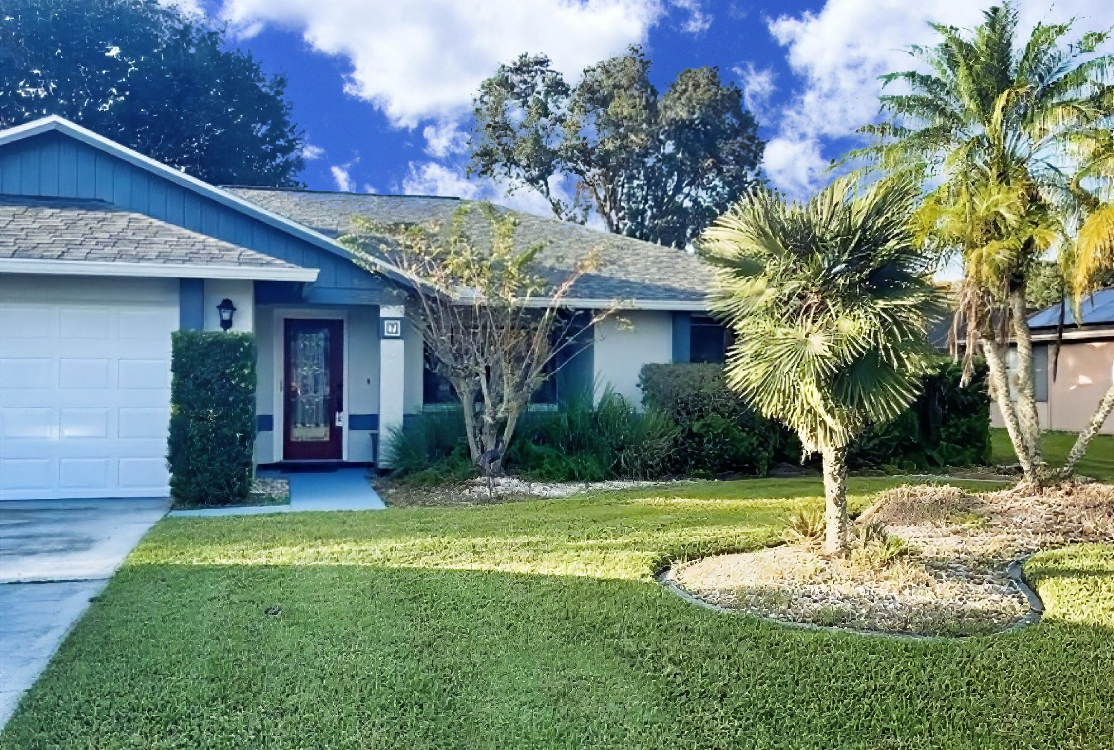 A single-story house with a blue garage, a red door, and a well-maintained lawn with a palm tree.