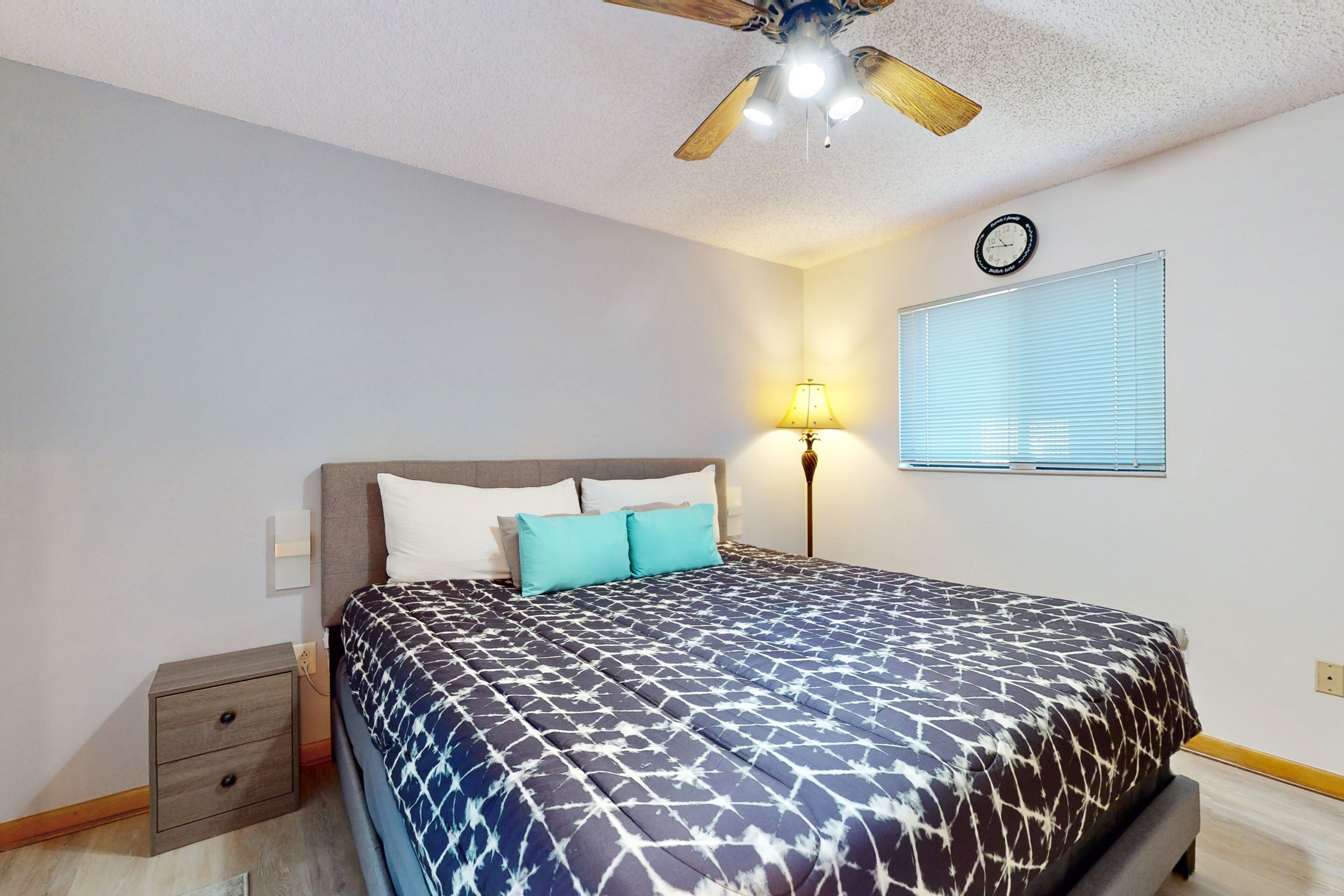 A bedroom with a bed, a nightstand, a lamp, a ceiling fan, and a window with blinds.