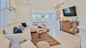 A spacious living room with a white sofa, a coffee table, and a TV mounted on the wall. - Bright 1940s Beach Cottage With Cabana Plus Private Pool (St. Augustine)