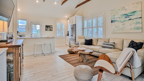 A spacious living area with a sofa, ottoman, and wooden coffee table. There is a kitchen area with a refrigerator and white cabinets. The room has large windows and a ceiling fan. - Bright 1940s Beach Cottage With Cabana Plus Private Pool (St. Augustine)