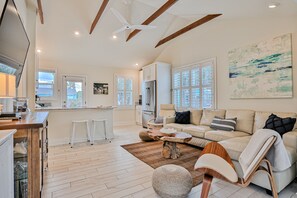 A spacious living area with a sofa, ottoman, and wooden coffee table. There is a kitchen area with a refrigerator and white cabinets. The room has large windows and a ceiling fan. - Bright 1940s Beach Cottage With Cabana Plus Private Pool (St. Augustine)