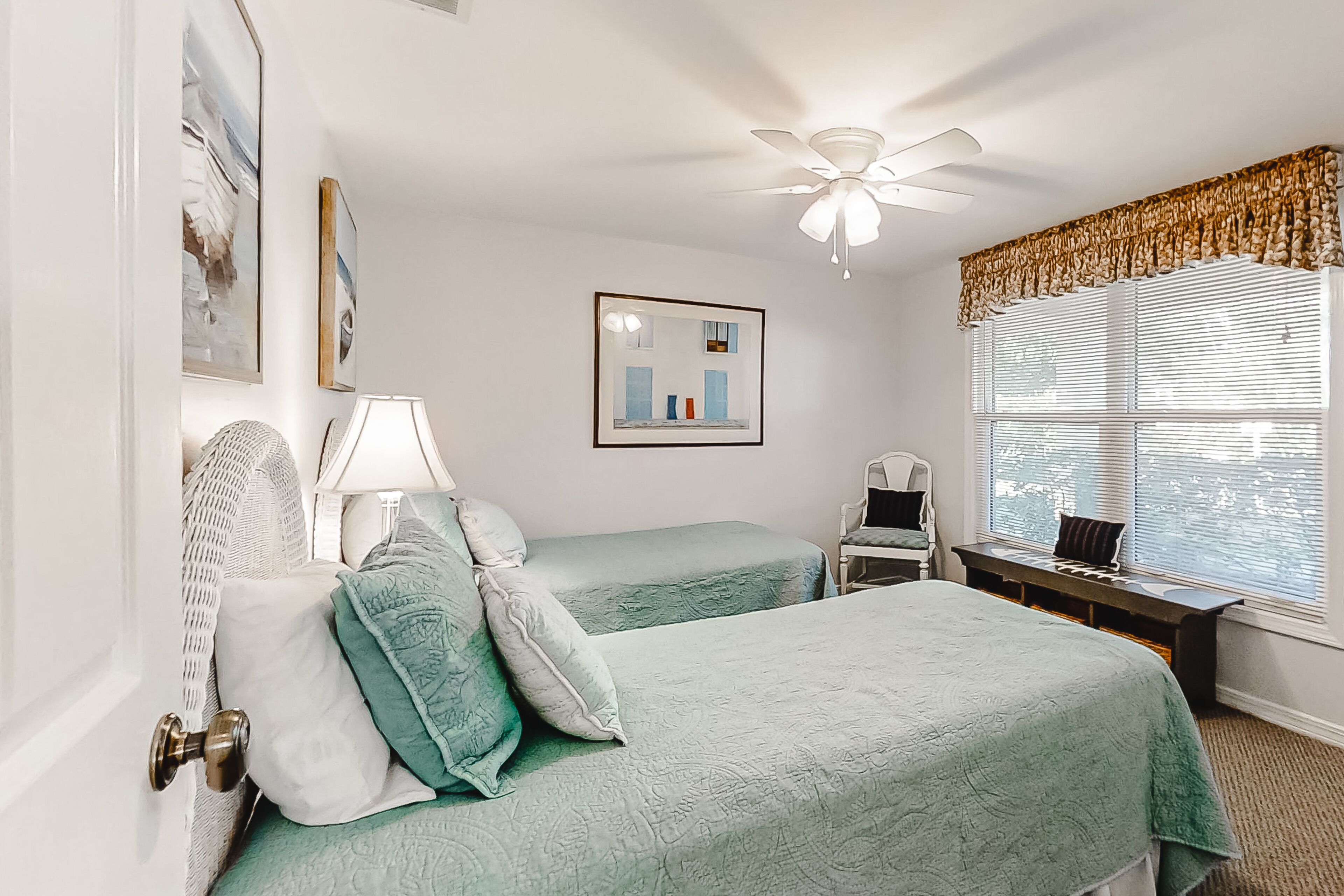A room with two beds, a ceiling fan, a window with blinds, a desk with a chair, and a lamp.
