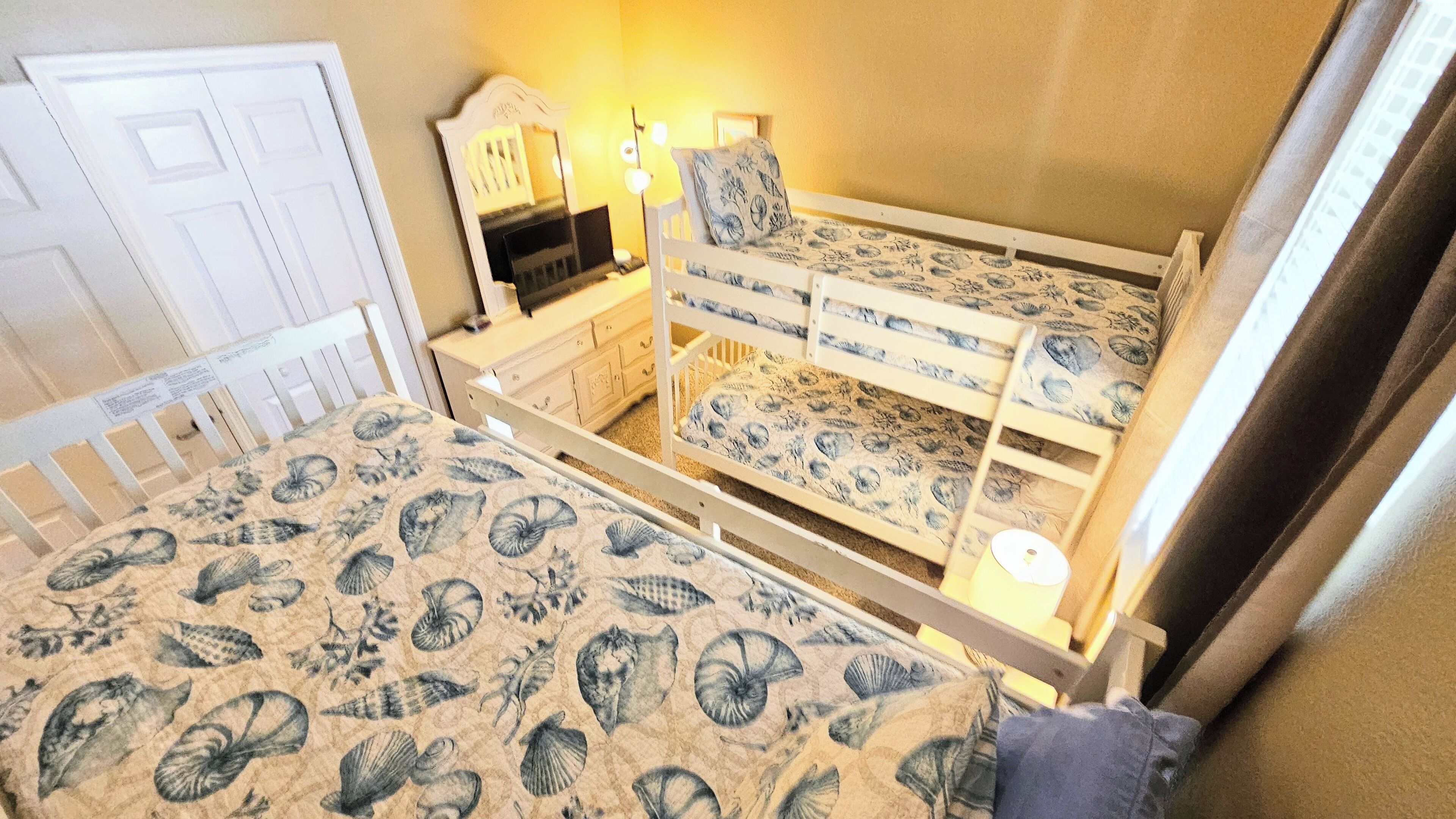 A bunk bed with a nautical-themed bedspread, a bedside table with a lamp, and a dresser with a mirror.