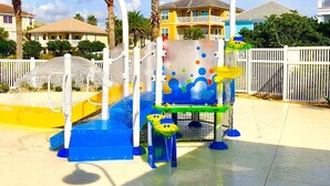 Un espace de jeux aquatiques avec des toboggans et des zones d’éclaboussures, entouré de palmiers et délimité par une clôture blanche.