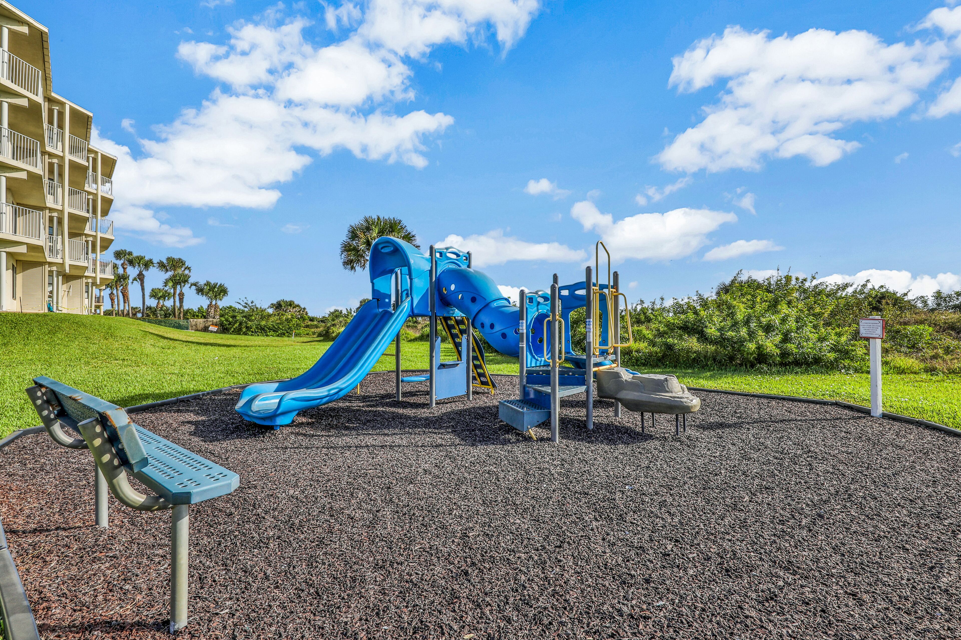 A playground with a blue slide and climbing structures, surrounded by a landscaped area with palm trees and a building in the background.