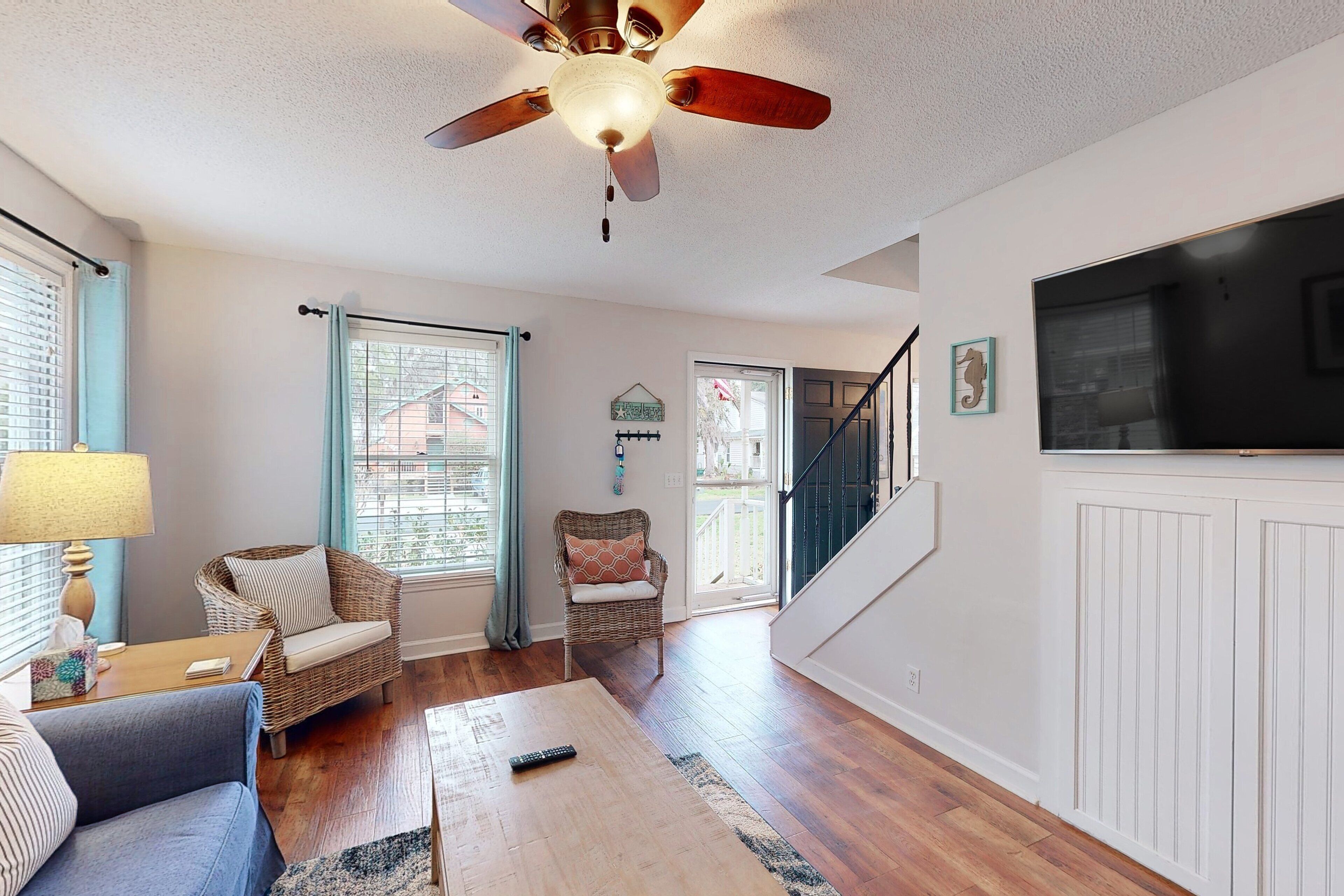 A living room with a ceiling fan, wicker furniture, a wooden coffee table, and a TV mounted on a white panelled wall.