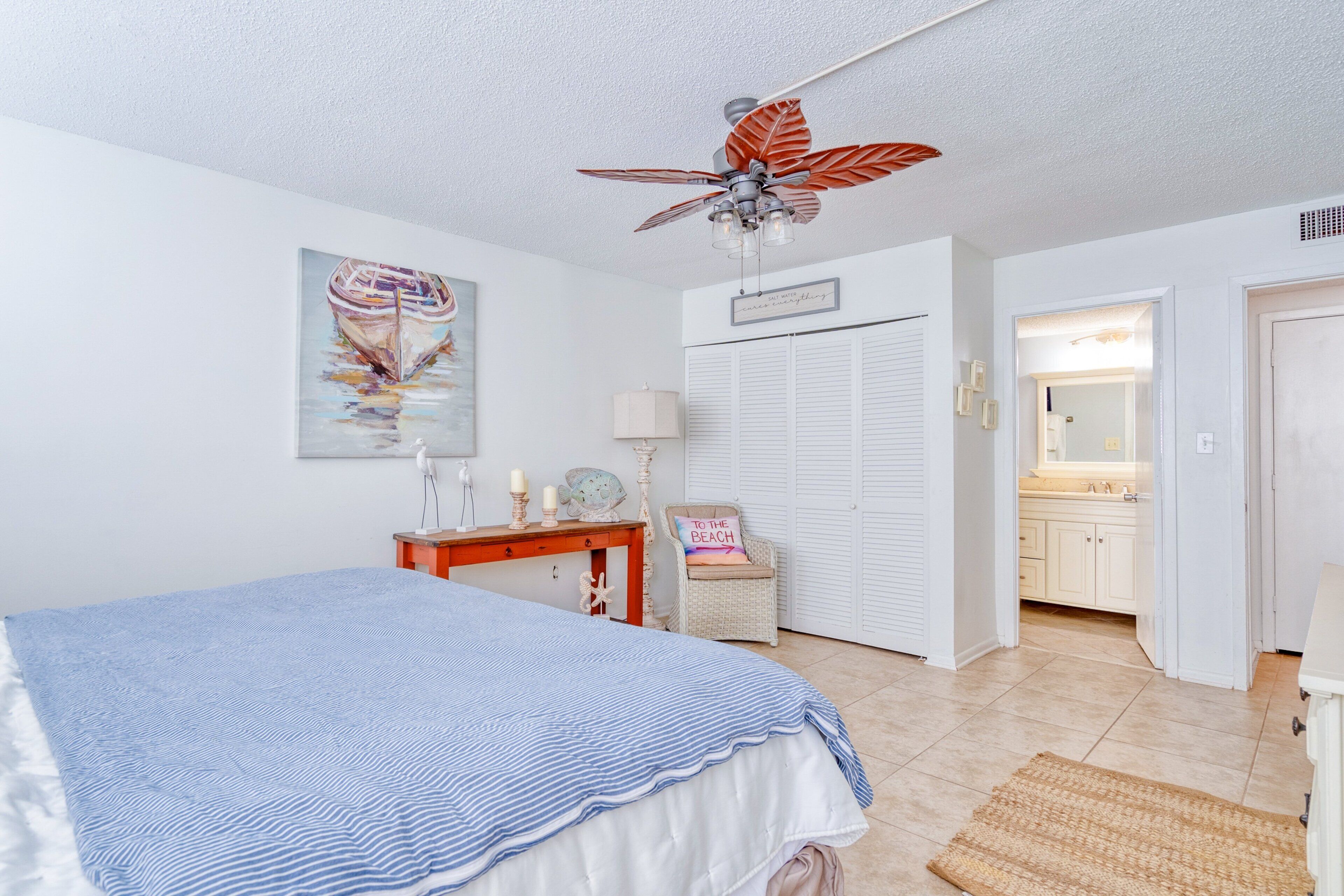 A bedroom with a bed, a desk, a chair, a ceiling fan, a painting, a wardrobe, and a bathroom visible through an open door.