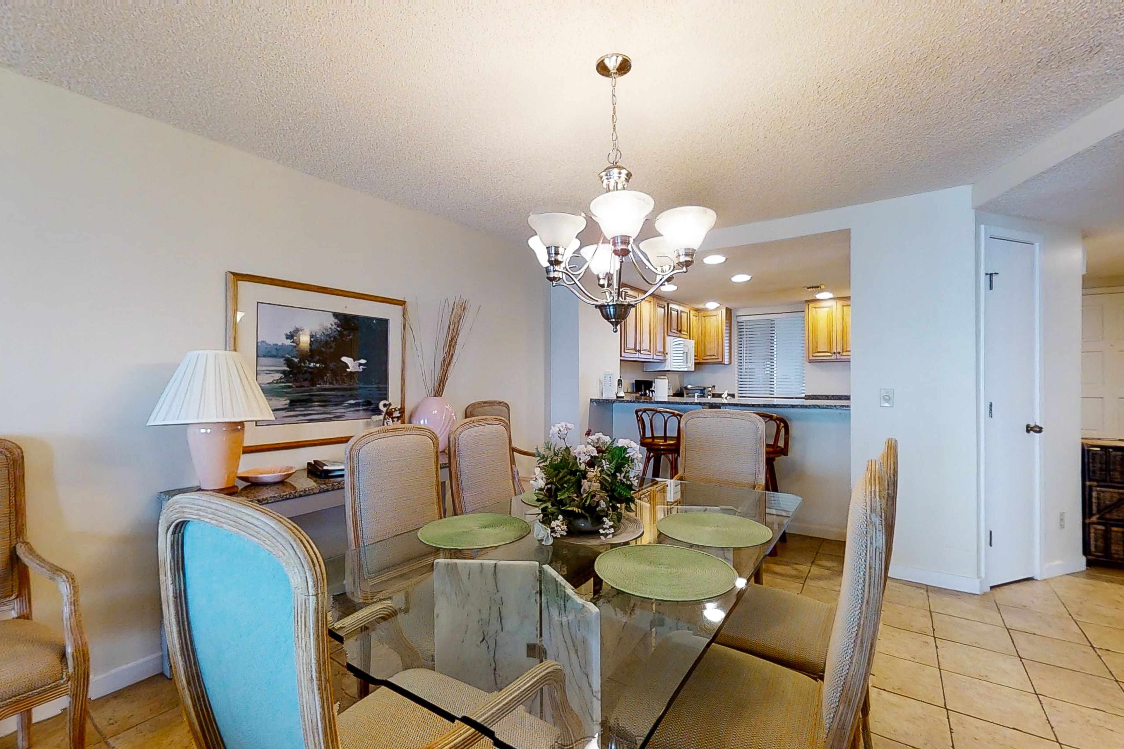 A dining area with a glass table, chairs, a chandelier, and a kitchen in the background.