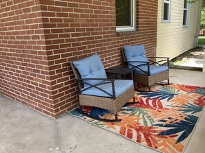 A patio area with two wicker chairs with blue cushions, a small table, and a colorful rug. - Classy Cottage With a Washer/dryer, Central AC, & Fast Wifi - Near Beaches (St. Augustine)