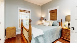 A bedroom with a wooden bed frame, a blue bedspread, two bedside lamps, a nightstand, and a wicker chest of drawers. - Classy Cottage With a Washer/dryer, Central AC, & Fast Wifi - Near Beaches (St. Augustine)