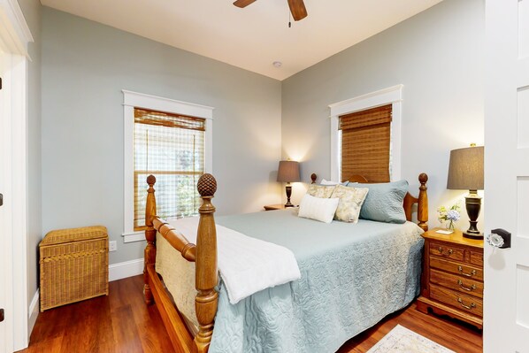 A bedroom with a wooden bed frame, a blue bedspread, two bedside lamps, a nightstand, and a wicker chest of drawers. - Classy Cottage With a Washer/dryer, Central AC, & Fast Wifi - Near Beaches (St. Augustine)