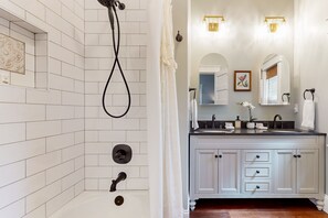 Uma casa de banho com chuveiro revestido a azulejos brancos, toucador duplo com armários e um espelho sobre o lavatório.
