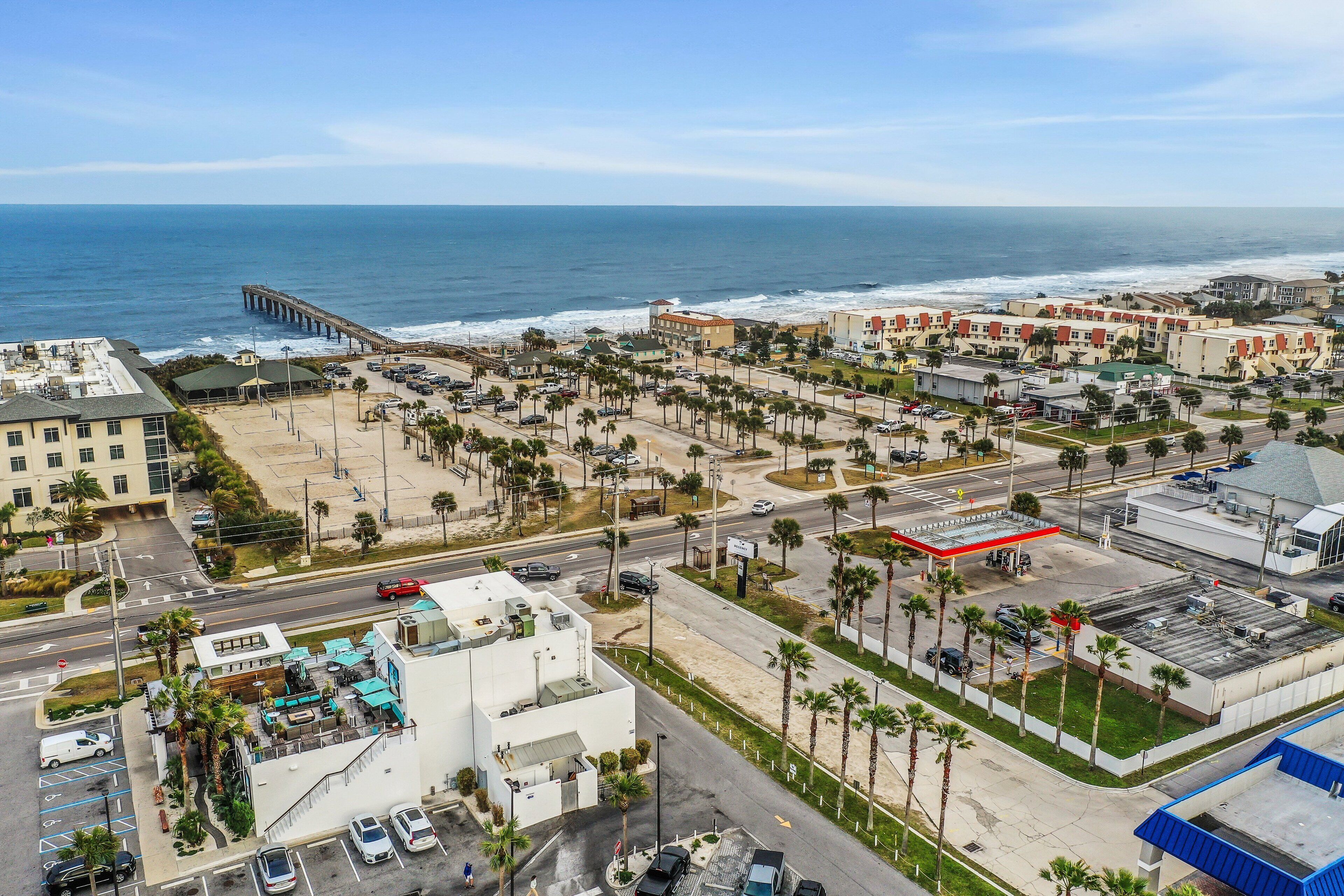 A coastal cityscape with a prominent pier extending into the ocean, a parking lot, and a row of buildings with a red-roofed gas station.
