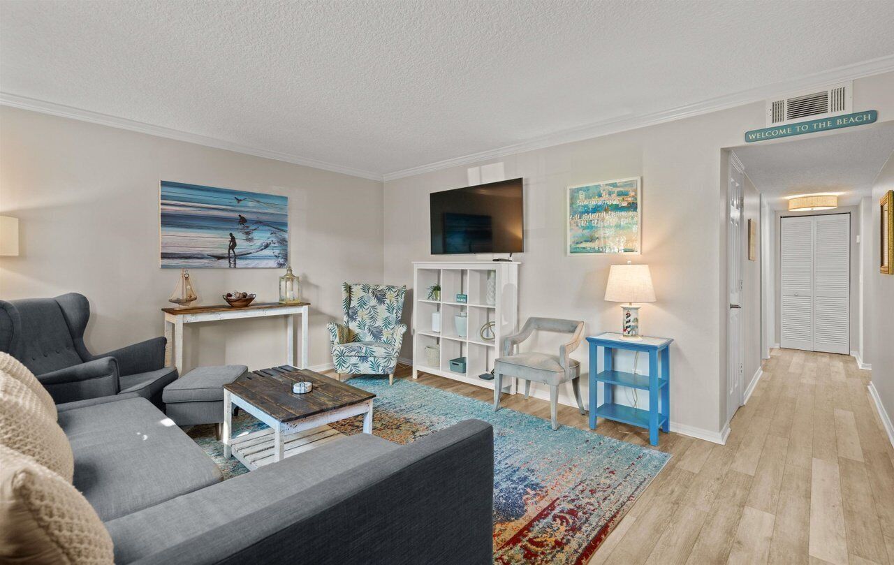 A modern living room with a grey sofa, a coffee table, a TV, and a bookshelf. There is a 'Welcome to the Beach' sign on the wall.