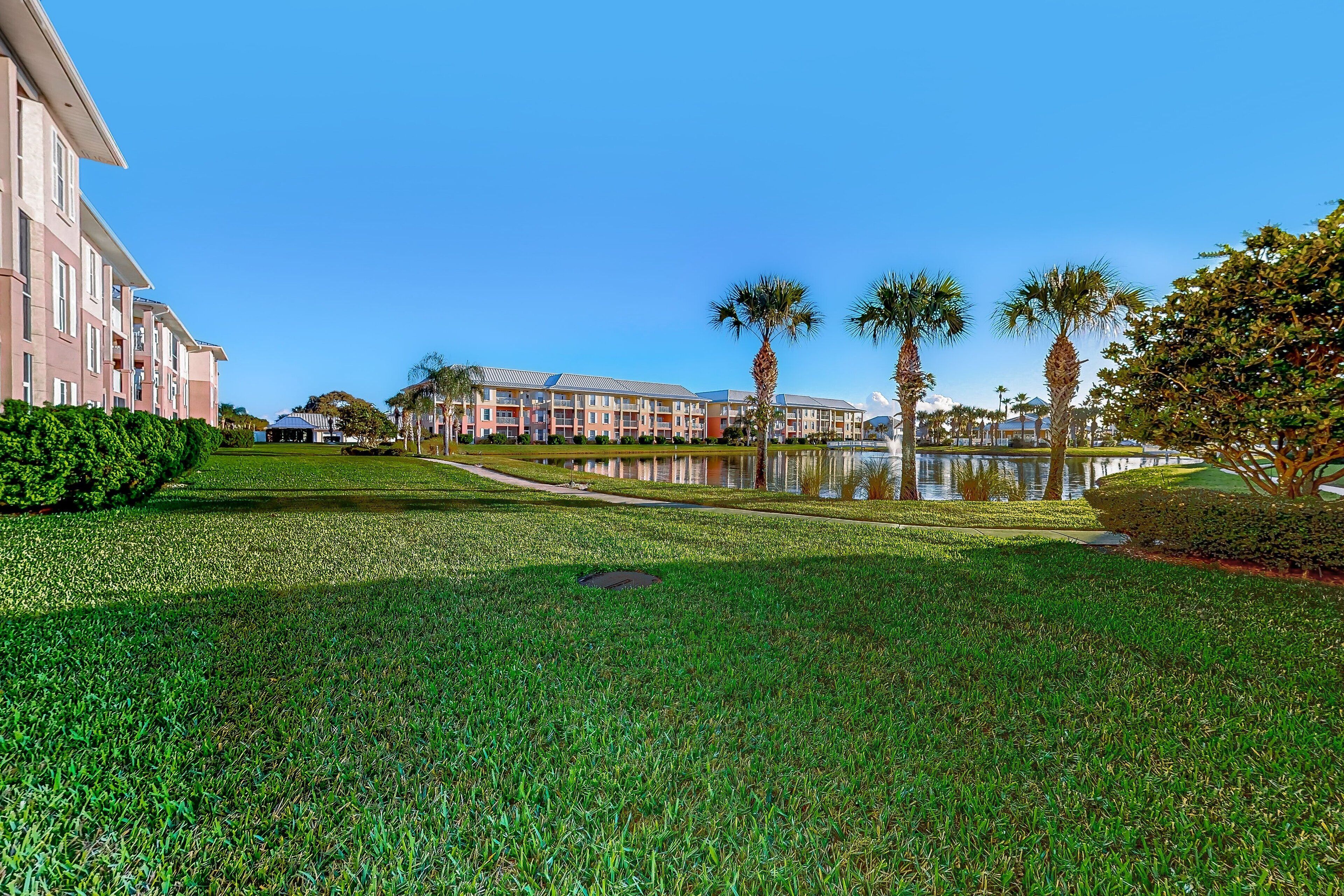 A hotel with a large lawn, palm trees, and a body of water reflecting the buildings.
