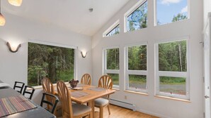 A dining area with a wooden table and chairs, large windows with a forest view, and a ceiling with recessed lighting.