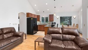 A modern living room with leather sofas, a wooden coffee table, and a kitchen with wooden cabinets and a black refrigerator.