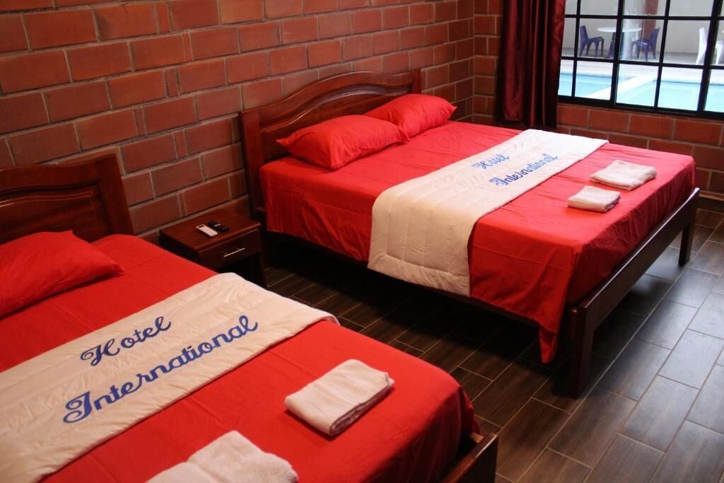 Two beds with red bedding and a white blanket, a wooden headboard, a brick wall, and a window with a view of outdoor furniture.