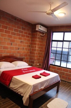 A hotel room with a brick wall, a bed with white bedding and red accents, a ceiling fan, and a window with a view of the outside. - Hotel International (Pedernales)