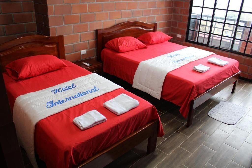 Two beds with red bedding and white towels, a brick wall, and a window with a view.