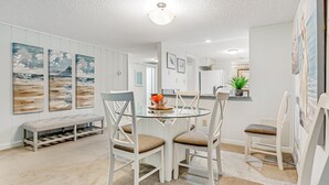 A dining area with a glass table, white chairs, and a bench. There are three paintings on the wall and a plant on the counter. - Beachwood Residence Nestled in a Canopy of Oak Trees (Fernandina Beach)