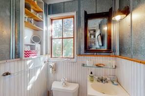 A bathroom with a toilet, sink, and a window. There are shelves with toiletries and a mirror above the sink. - Charming Getaway w/ a Kitchenette & Free Wifi - Walk to the Beach (St. Simons Island)