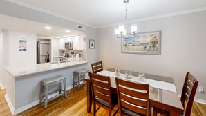 A modern kitchen with white cabinetry, a central island with stools, and a dining area with wooden chairs and a white table. - Beach Wood Wonder With Pool Access - Walk to Ocean (Fernandina Beach)