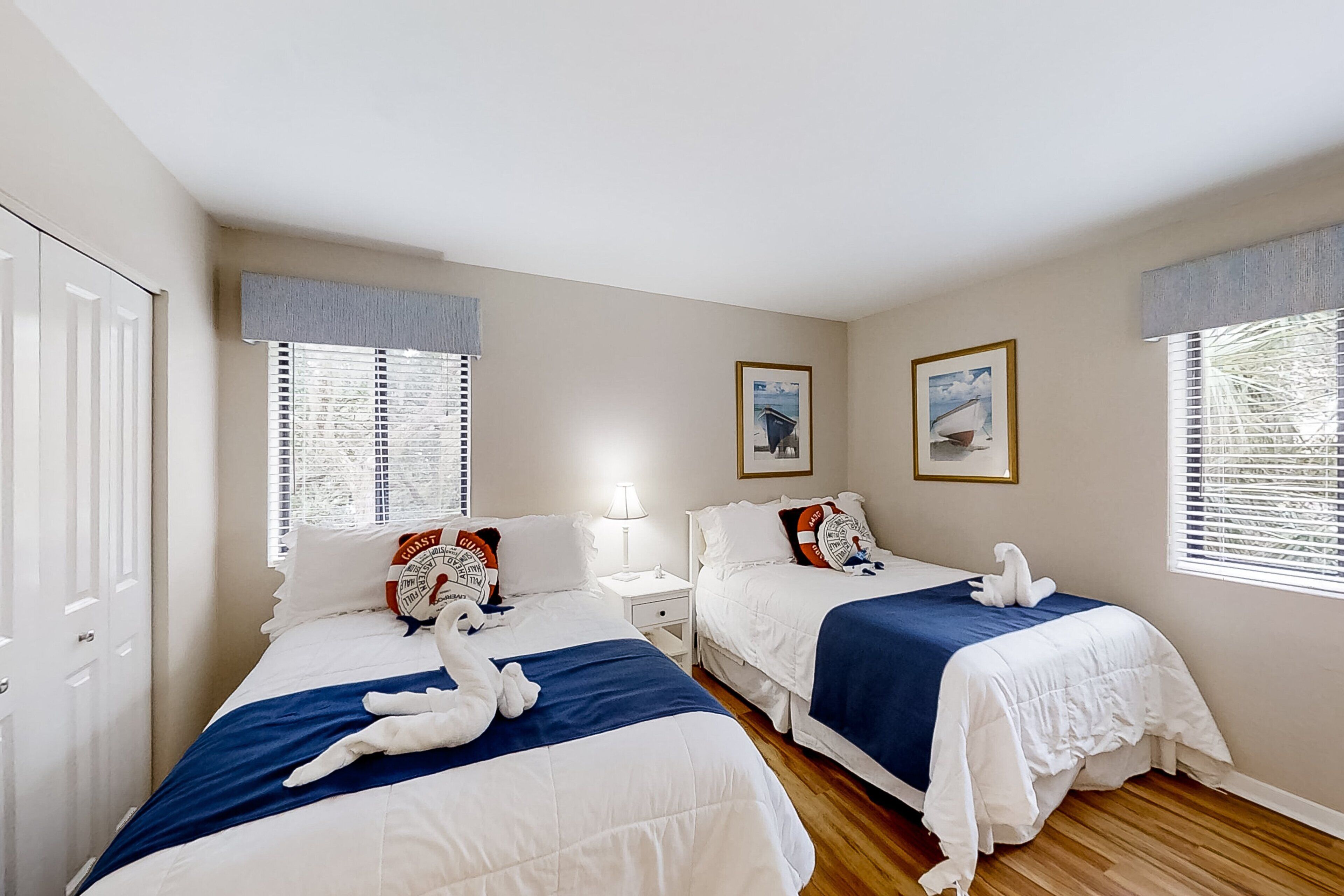 A hotel room with two beds, each with a blue blanket and white pillows, a nightstand with a lamp, and a framed picture on the wall.