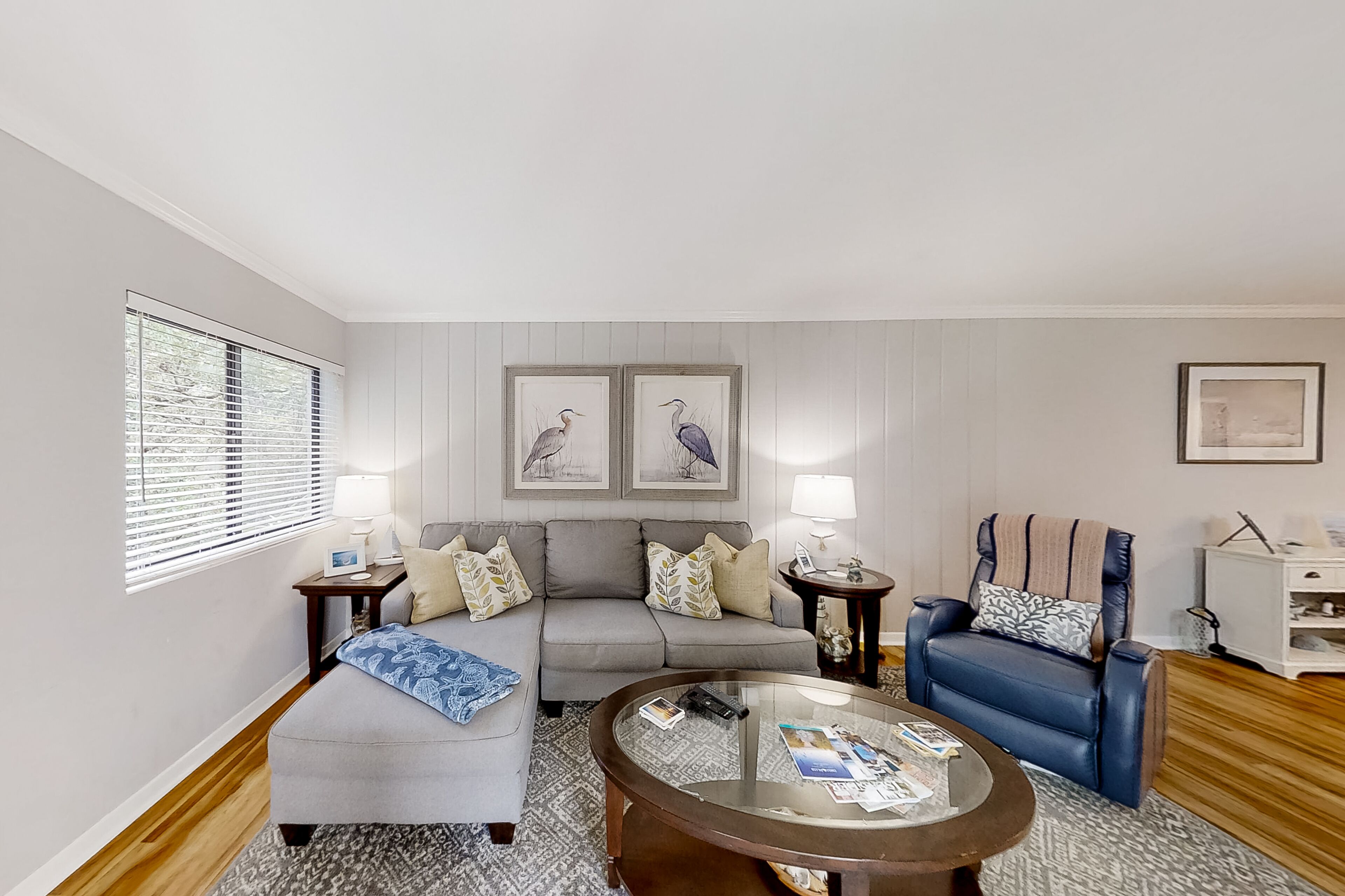 A living room with a grey sofa, a blue armchair, a glass coffee table, and a patterned rug. There are two side tables with lamps and framed artwork on the wall.
