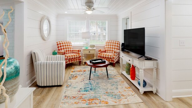 Una sala de estar con televisor, dos sillones estampados, un sofá a rayas, una mesita de centro y un jarrón decorativo.