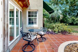 Une terrasse extérieure avec un sol carrelé, une petite table ronde accompagnée d’une chaise, et une fenêtre donnant sur le jardin.