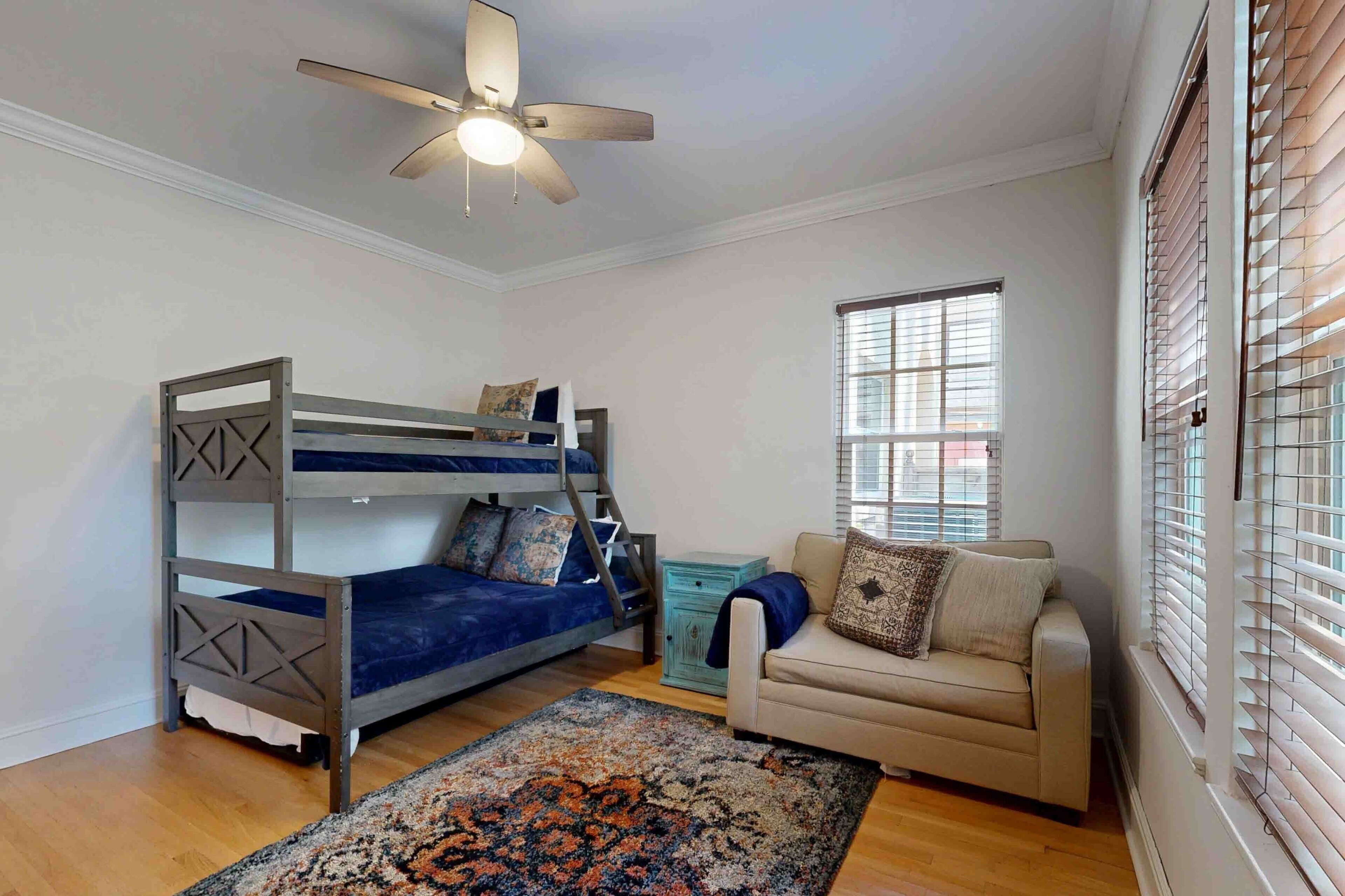 A room with a bunk bed, a sofa, a ceiling fan, and a window with blinds.