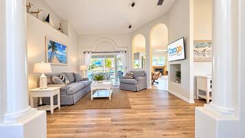 A spacious living room with a grey sofa, a white coffee table, and a view of the outdoors through large windows.