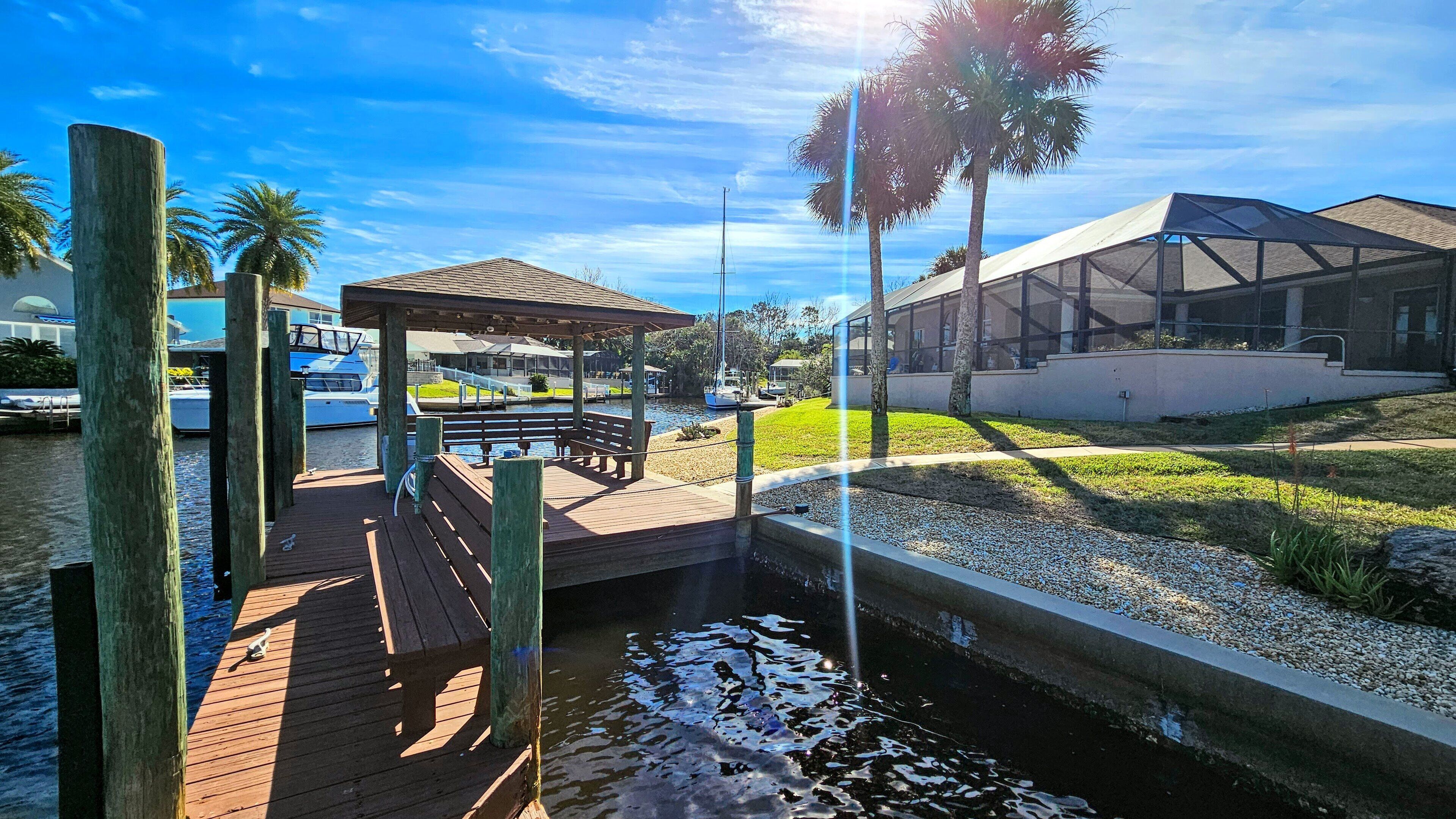 A wooden dock with a gazebo and benches, a marina with boats, and a modern building with large windows.