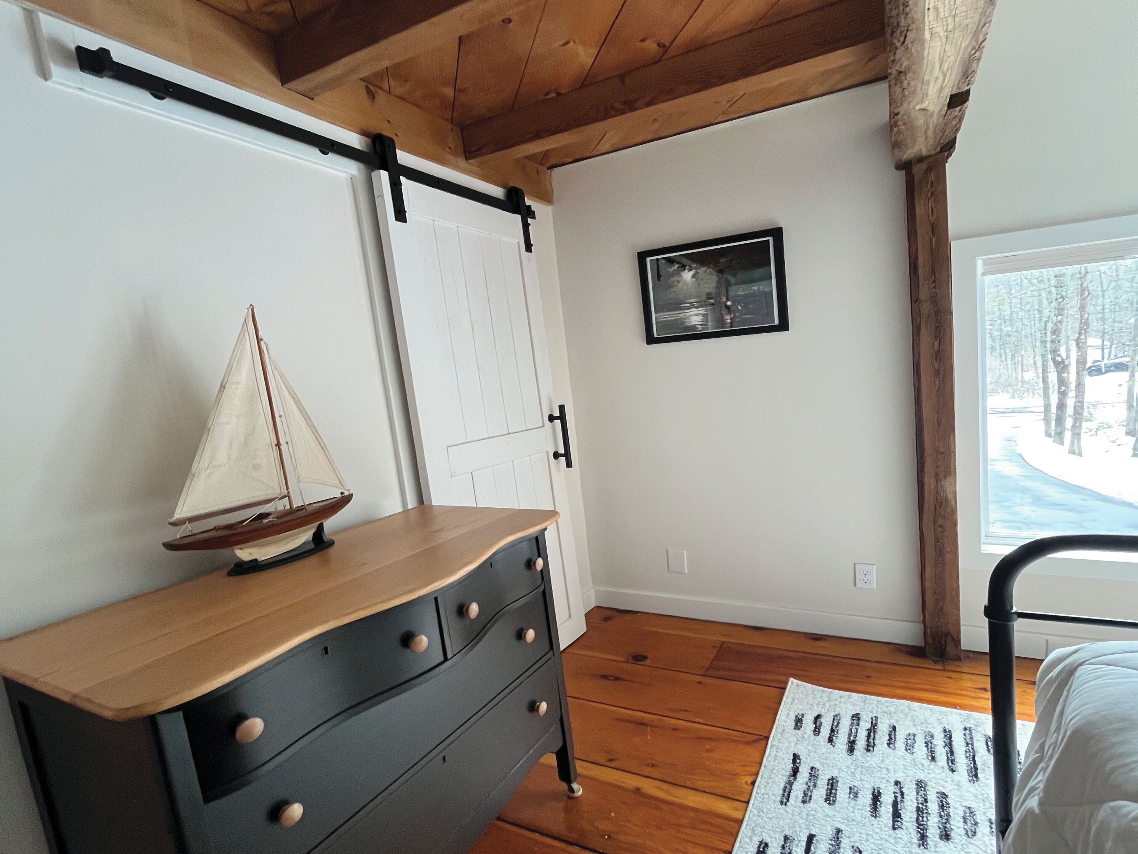 A room with a wooden dresser, a sailboat model, a framed picture, and a window with a view of trees.