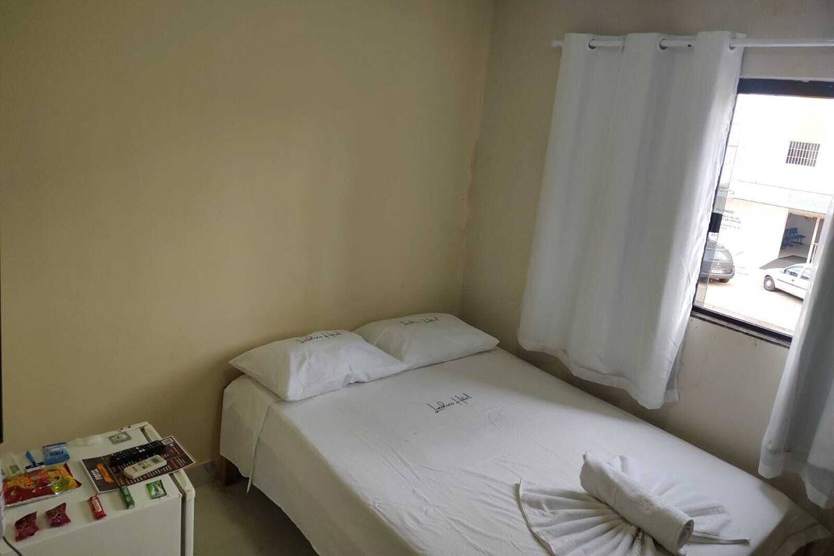 Habitación individual con una pared sencilla, una ventana con cortinas blancas y una mesita de noche con artículos de aseo.