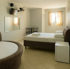 Ein Hotelzimmer mit einem Bett, einem kleinen Tisch mit Stühlen, einem Kühlschrank, einem großen, runden Sessel, einem Fernseher und einem Fenster mit Vorhängen.