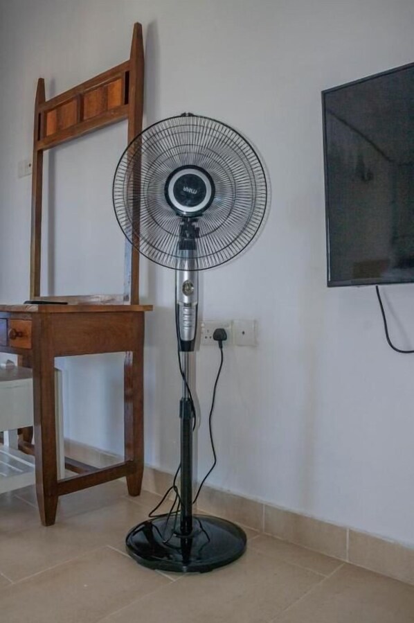 Un ventilador de pie con una rejilla circular, una silla de madera y un televisor montado en la pared.