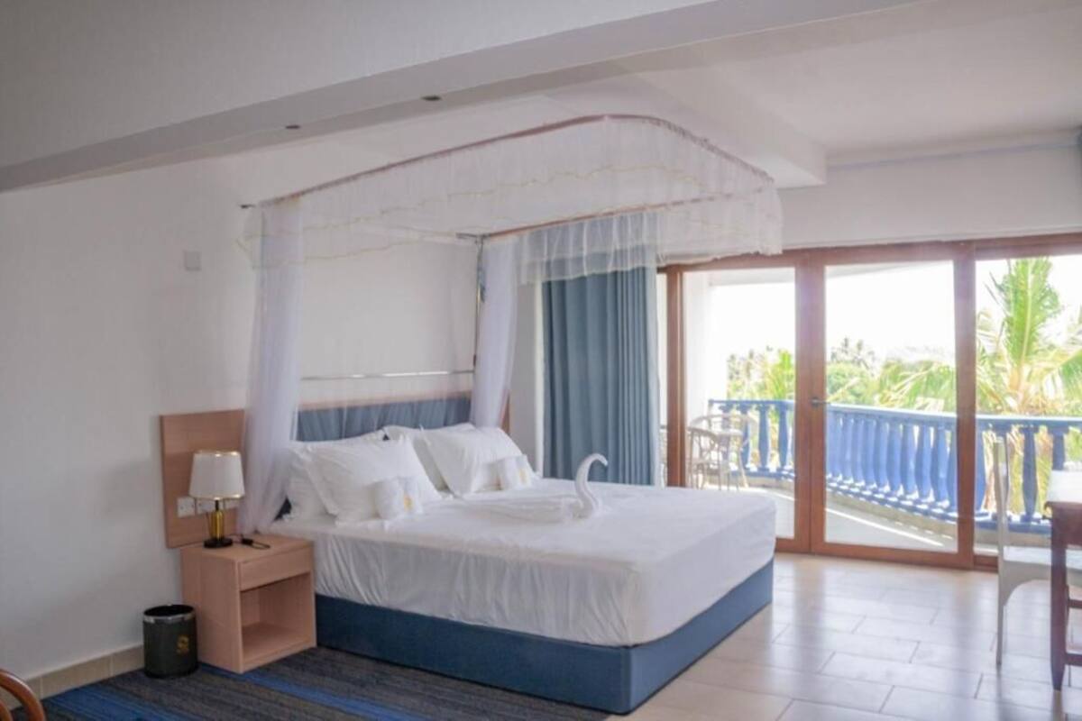 A hotel room with a bed, a nightstand, a small trash bin, and a balcony with a view of greenery.