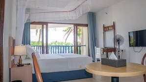 A hotel room with a bed, a bedside table with a lamp, a round table with a tissue box, a fan, a TV, and a balcony with a view of greenery. - GSIH Hotel Diani (Diani Beach)