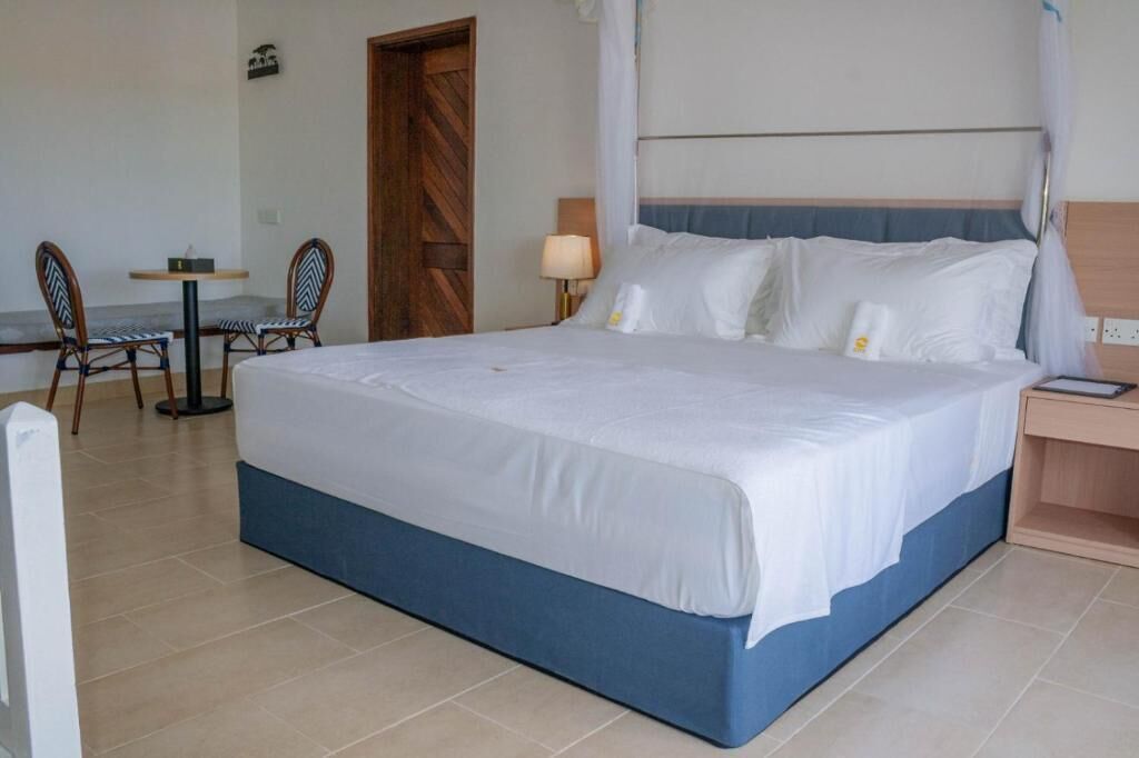 A hotel room with a large bed, a bedside table, a chair, and a small table with a lamp.