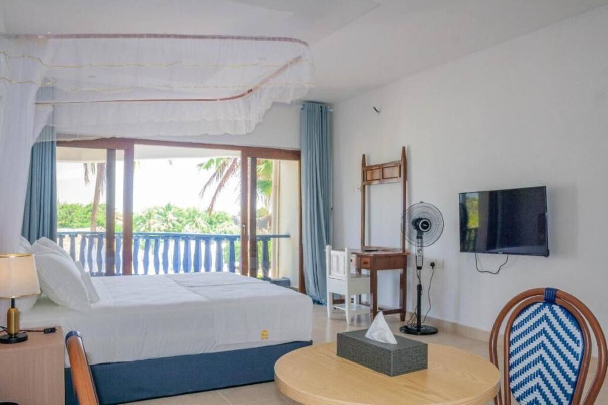A hotel room with a bed, a desk, a chair, a TV, and a balcony with a view of palm trees.