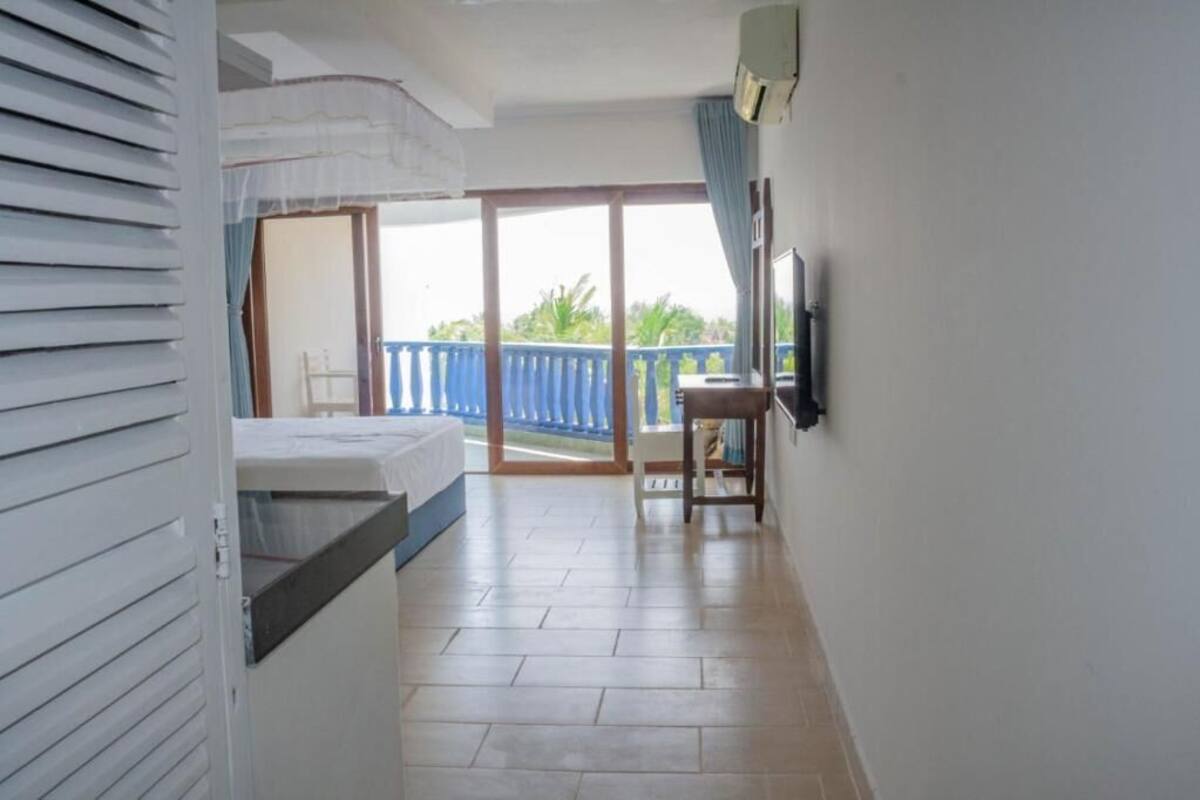 A room with a bed, a small table, a TV mounted on the wall, and a balcony with a blue railing.