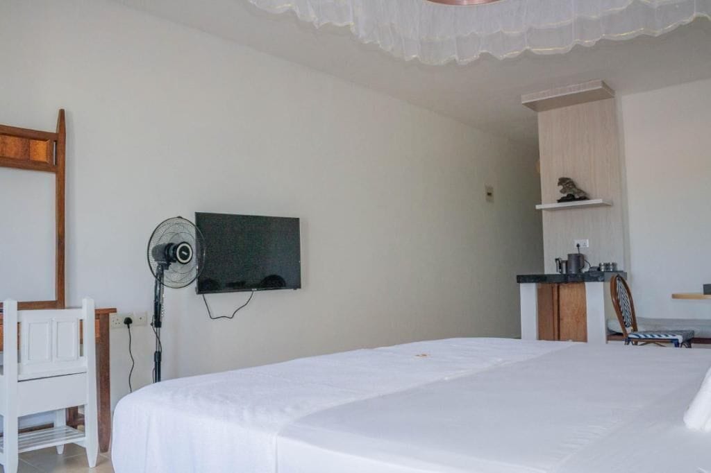 A bedroom with a bed, a TV mounted on the wall, a fan, a small kitchenette, and a chair.