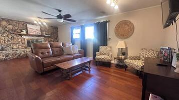 Una sala de estar con pared de piedra, un sofá de cuero marrón, un sillón estampado, una mesa de centro de madera y un ventilador de techo.