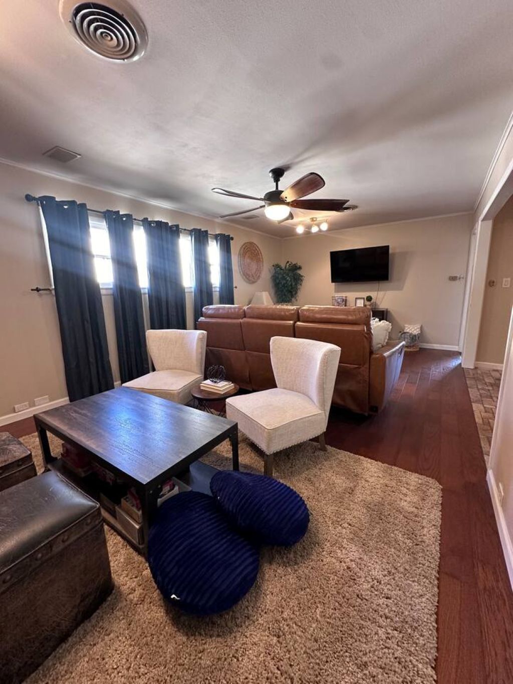 Un salon avec une table en bois foncé, deux fauteuils blancs, un canapé marron, un ventilateur de plafond et un téléviseur fixé au mur.